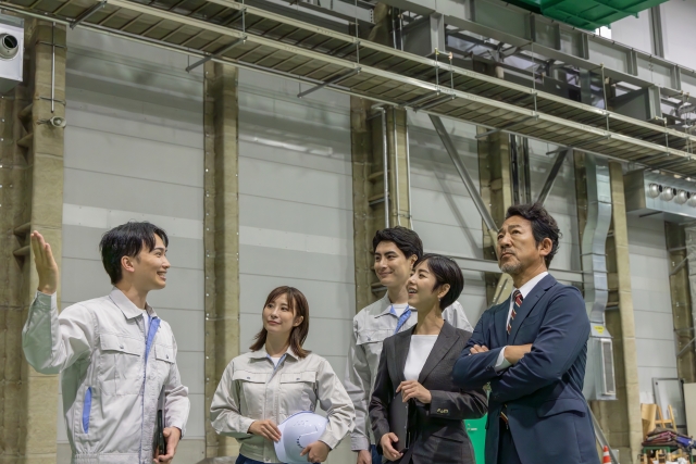 工場内で打ち合わせを行う作業服姿の技術者とスーツ姿のビジネスマンたち