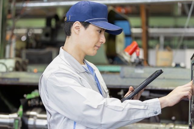工場内で作業着とキャップを着用した男性作業員がタブレットを持ちながら設備を操作している様子